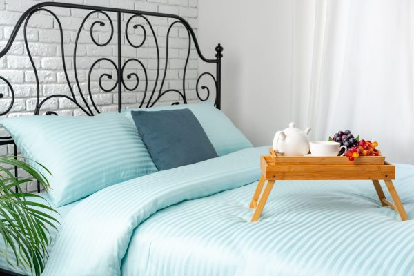A wooden breakfast tray sits on a blue bed, containing a teapot, grapes, and a cup. The tray is folded out, suggesting that it is being used.